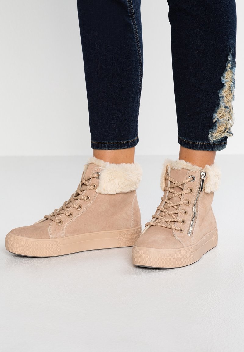 Baskets montantes beige avec bordure en fourrure et fermeture éclair latérale portées avec un jean skinny déchiré foncé sur une surface claire et plate.