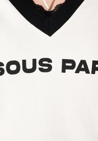 Witte T-shirt met een zwarte V-hals en kanten rand, met vetgedrukte zwarte tekst "SOUS PAR" aan de voorkant.