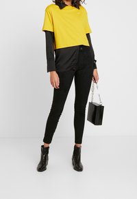 Gul kortärmad t-shirt med svart krage, lager på lager över en svart långärmad skjorta. Svarta skinny jeans och svarta ankelstövlar, håller en svart handväska.