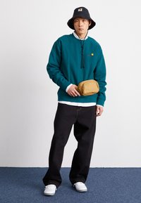 Teal genser med snor, hvit skjorte med krage under, svart bøttehatt, svarte bukser og en gyllen fanny pack.