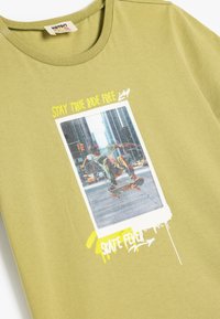Camiseta de algodón verde oliva con un estampado gráfico de un skater, enmarcado por salpicaduras coloridas y el texto: "MANTENTE VERDADERO RÍE LIBRE" y "FIEBRE DEL SKATE".