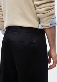 Homme portant un pull beige par-dessus une chemise rayée bleue rentrée dans un pantalon noir sur mesure, vu de la taille vers le bas, de dos.