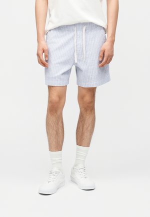 Muž v svetlomodrých prúžkovaných šortkách so sťahovacou šnúrkou, bielych ponožkách a bielych teniskách, stojaci so zvesenými rukami.