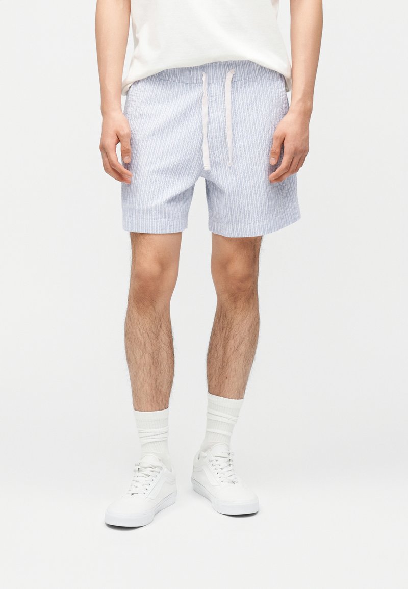 Άνδρας που φοράει ανοιχτό μπλε ριγέ σορτς με κορδόνι, λευκές κάλτσες και λευκά αθλητικά παπούτσια, στέκεται με τα χέρια χαλαρά δίπλα στο σώμα.