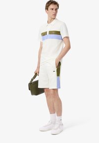 Polo blanc avec des blocs de couleurs vert, bleu et lilas. Associé à un short blanc avec un accent vert et un sac à épaule vert.