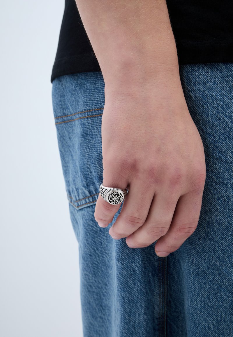 Anel de prata com intrincadas gravuras a preto, usado numa mão. O fundo apresenta tecido de ganga azul com costura e textura visíveis.
