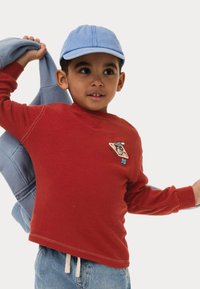 Rood langarm shirt met geborduurd graphic, denim shorts en een lichtblauwe pet die met een grijze sweatshirt over de schouder hangt.