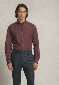 Camisa de botões cor castanho-avermelhado com colarinho, usada com calças cinzentas de cintura alta. Tecido liso, corte justo, com botões frontais e mangas compridas.