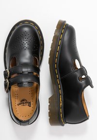 Sorte lær sko med utskjæringer, to spenner, gult stikningsdetalj, og en kraftig brun såle. Perforert overdel for ventilasjon.