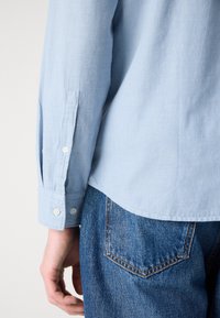 Bras gauche et main d'une personne portant une chemise à manches longues bleu clair et un jean en denim bleu sur un fond uni.