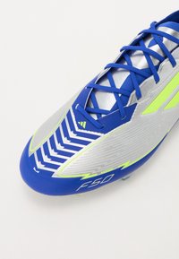 Cizme de fotbal albastre și argintii, cu un superior texturat și modele în zigzag, șireturi albastre și accente verzi. Modelul este denumit "F50."