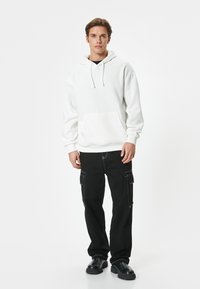 Felpa bianca con cappuccio e tasca frontale, abbinata a pantaloni cargo neri con tasche laterali e dettagli in cucitura bianca. Calzature nere.