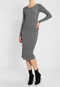 Abito grigio a maglia con maniche lunghe, texture a coste, scollo rotondo e spacco laterale. Abbinato a stivaletti neri con tacco alto.
