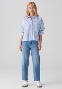 Camicia azzurro chiaro a righe con taschino sul petto, abbinata a jeans azzurro chiaro e sneakers bianche. Tessuto morbido, vestibilità comoda.