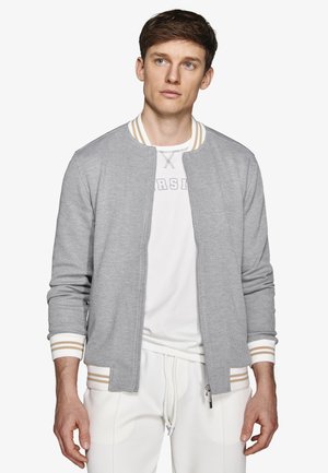 Junger Mann mit einer hellgrauen, mit einem Reißverschluss ausgestatteten Strickjacke mit beigen gestreiften Bündchen und Kragen, die über einem weißen Hemd und weißen Hosen getragen wird.