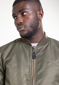 Olijfgroene bomberjas met een ritssluiting aan de voorkant, geribbelde kraag en tonale stiksels. Voorzien van een merklogo op de borst. Soepele textuur.