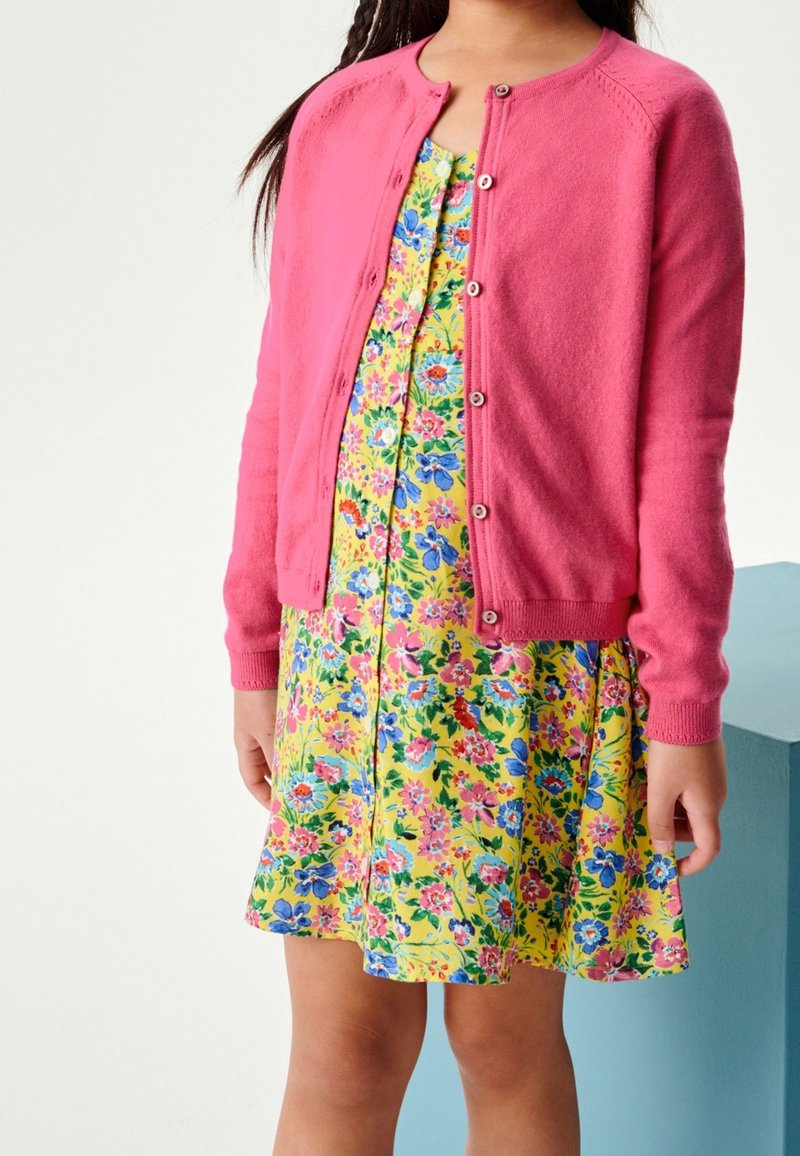 Enfant portant un cardigan boutonnière rose vif sur une robe jaune à fleurs bleues, roses et rouges, debout devant un fond uni.