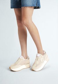 Sneakers atletici beige con materiali in rete e suede, punta rotonda, suola intermedia bianca e suola testurizzata. Presentano una chiusura con lacci e un dettaglio di branding.