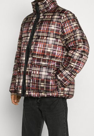Homme portant une doudoune à carreaux marron, rouge et crème avec de grandes poches avant et un jean noir, debout devant un fond blanc.