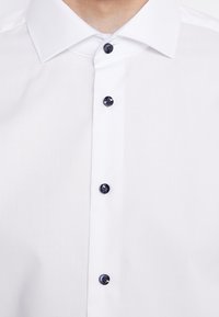 Zbliżenie białej koszuli z czterema zapiętymi ciemnymi guzikami i klasycznym, szpiczastym kołnierzem widocznym na osobie.