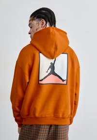 Haut à capuche orange en coton avec un grand logo Jordan graphique au dos, présentant une silhouette noire sur fond blanc.