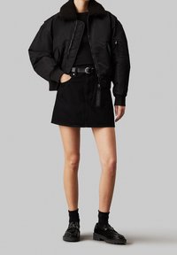 Giacca bomber nera con colletto in pelliccia sintetica, abbinata a una mini gonna in denim nera. Calze nere alla caviglia e scarpe lucide completano l'outfit.