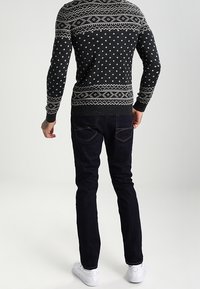 Maglione nero lavorato a maglia con motivi geometrici bianchi, abbinato a jeans scuri. Sneakers bianche completano l'outfit. Cuciture visibili sui jeans.