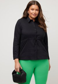 Camicia nera a maniche lunghe con bottone frontale e vita con cordino, abbinata a pantaloni verdi e una borsa nera.