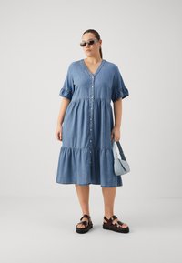 Denimdress med V-hals, korte volangærmer, knaplukning foran og lagdelt kant. Båret med sorte sandaler og en lyseblå crossover-veske.
