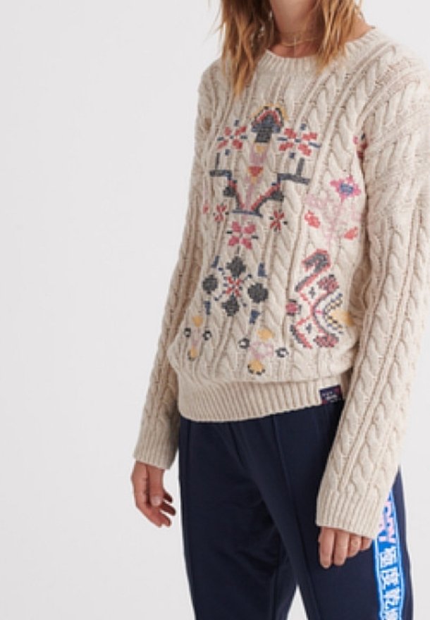 Femme portant un pull en maille torsadée crème avec une broderie florale colorée, associé à un pantalon bleu marine avec une bande latérale bleue.