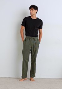 Pantalons rayés verts avec une taille à cordon de serrage et des poches latérales, associés à un t-shirt noir ajusté. Le mannequin se tient pieds nus contre un fond uni.
