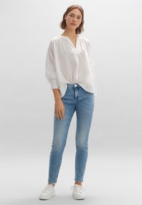 Blouse blanche à manches longues avec un décolleté en V et une coupe ample, associée à un jean slim bleu et des baskets blanches sur un fond gris clair.