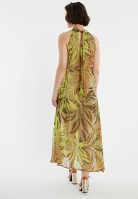 Robe maxi en tissu léger et transparent avec un motif de feuilles vertes et brunes. Présente un col halter et un détail de nouage à l'arrière.