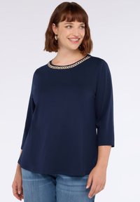 Frau mit kurzen braunen Haaren, die ein marineblaues Oberteil mit Kettendetail am Ausschnitt und hellblaue Jeans trägt. Sie lächelt und sieht nach rechts.