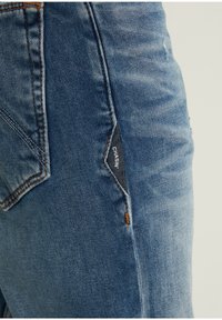 Modré džínové kalhoty s texturovaným povrchem, zdobené kontrastním oranžovým štepováním a černou látkovou visačkou s názvem značky.