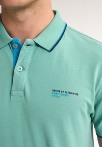 Svetlozelená polo košeľa vyrobená z textúrovaného materiálu, s tyrkysovým golierom a námorníckym akcentom, s potlačeným brandingom v modrej a čiernej farbe.