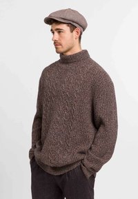 Chunky gebreide bruine trui met een hoge kraag, voorzien van een textuurpatroon. Gepaard met een lichtbruine platte pet.