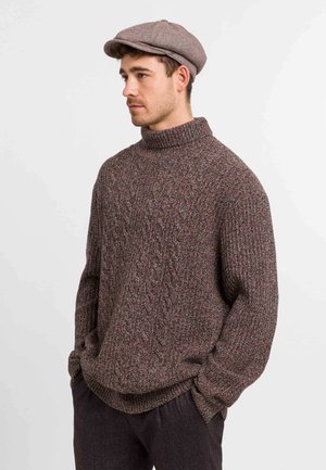 Chunky knit brown sweater with a high collar, featuring a textured pattern. Paired with a light brown flat cap.