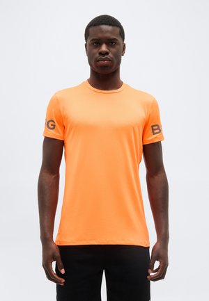 Άνδρας που στέκεται προς τα εμπρός φοράει φωτεινό πορτοκαλί κοντομάνικο πουκάμισο με μαύρα γράμματα "B" και "G" στα μανίκια και μαύρο παντελόνι.