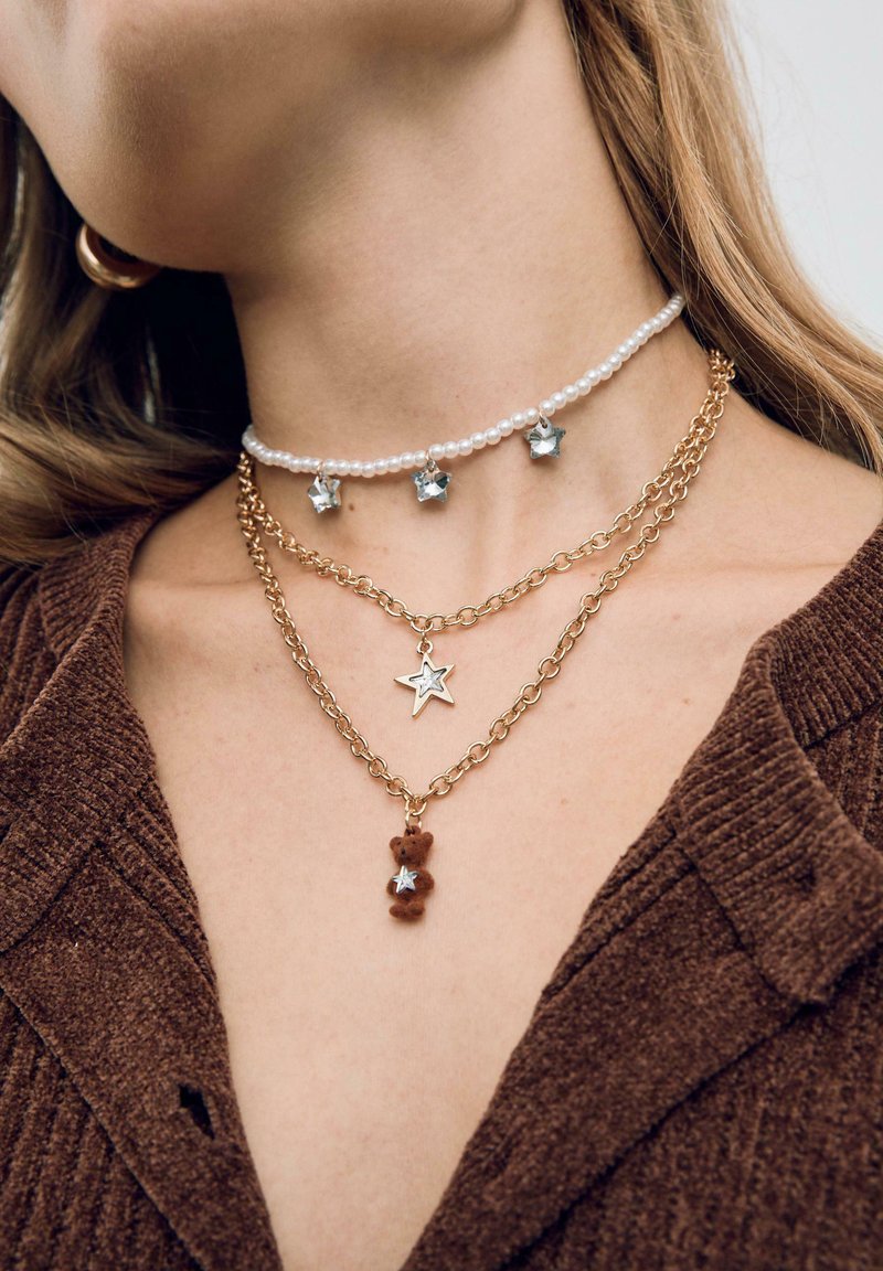 Collier ras du cou avec des perles rondes blanches et des accents en étoile argentés ; chaîne en or superposée avec un charme en étoile et un pendentif ours brun.