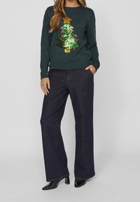 Grön tröja med en paljettdekorerad julgran, prydd med röda ornament och en gyllene stjärna. Bärs tillsammans med mörk denimbyxor med vida ben och lackskor.