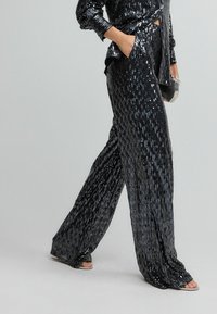 Schwarze Pailletten-Hosen im weit geschnittenen Design, mit einem glänzenden, strukturierten Finish, Seitentaschen und dezenten vertikalen Streifenmustern.