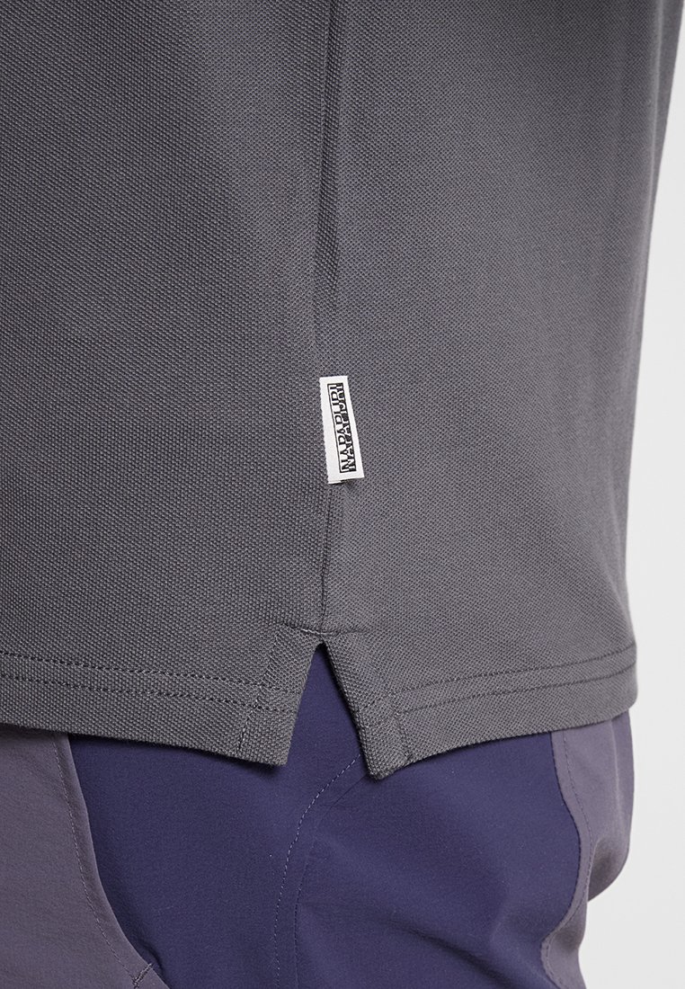 Nahaufnahme des grauen, strukturierten Poloshirt-Saums mit einem kleinen weißen Markentag oberhalb des seitlichen Schlitzes, getragen über einer dunkelblauen Hose.