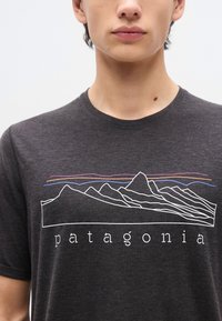 Νεαρό άτομο που φοράει σκουρόγκρι μπλουζάκι Patagonia με λευκό σχέδιο γραμμής βουνού και χρωματιστές οριζόντιες γραμμές στο στήθος.