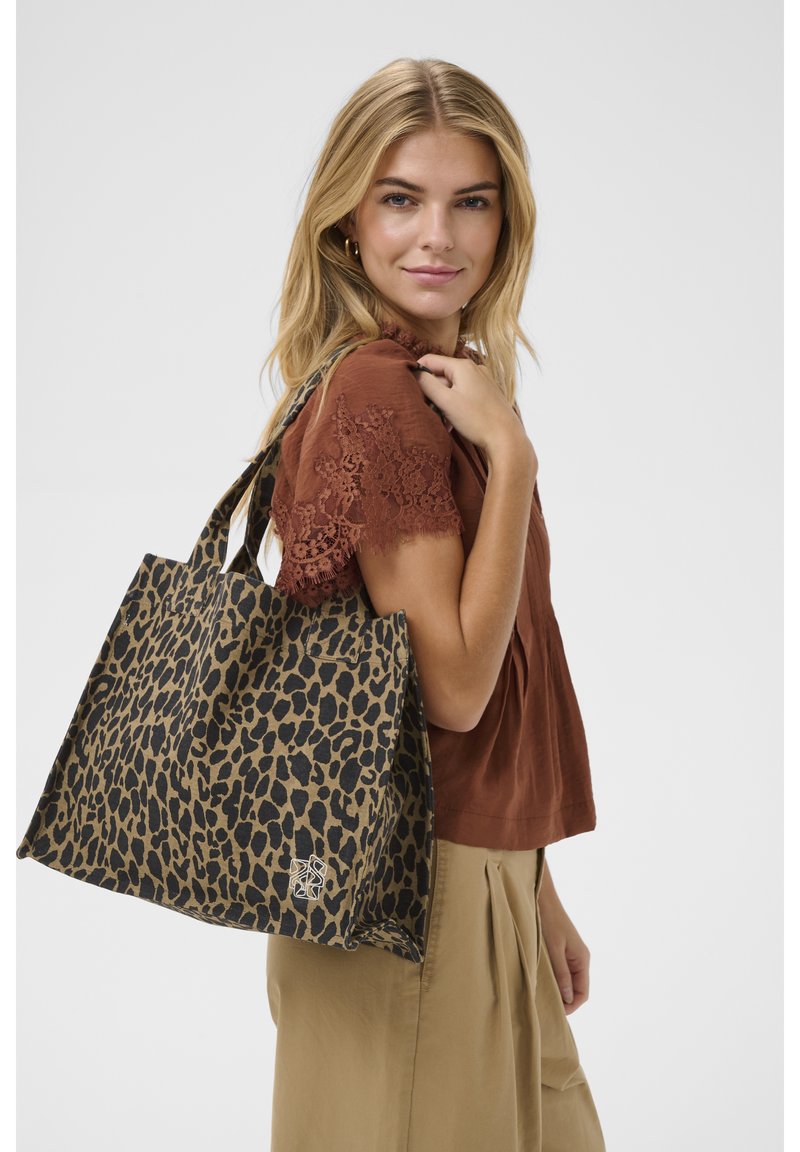 Femme blonde portant un haut à manches en dentelle marron et un pantalon beige, tenant un grand sac fourre-tout à imprimé léopard sur l'épaule, sur fond blanc.