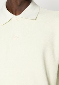 Svetle zelená polo triko vyrobené z texturované tkaniny, s klasickým límečkem a dvěma růžovými knoflíky na zapínání.