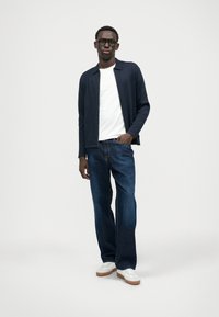 Ναυτικό πλεκτό cardigan πάνω από λευκό t-shirt, συνδυασμένο με σκούρο τζην παντελόνι και λευκά αθλητικά παπούτσια. Το μοντέλο στέκεται χαλαρά με τα χέρια στις τσέπες.