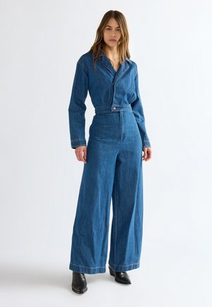 Frau trägt einen langärmeligen blauen Denim-Overall mit weitem Bein und schwarzen Knöchelstiefeln und steht vor einem schlichten weißen Hintergrund.