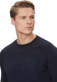 Felpa blu navy con scollo rotondo, maniche raglan e logo "GUESS" in rilievo sul davanti. Realizzata in tessuto morbido, design minimalista.