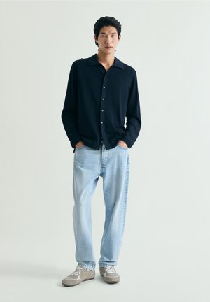 Joven de pie con las manos en los bolsillos, vistiendo una camisa oscura abotonada, vaqueros azul claro y zapatillas grises sobre un fondo liso.